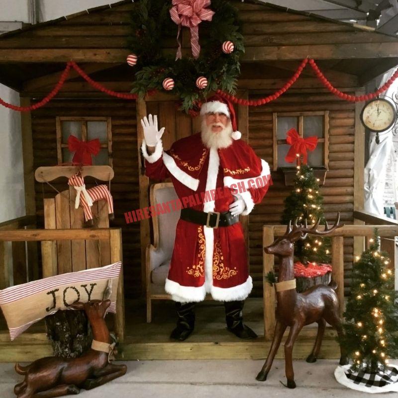 Real beard Santa for hire waving from a cabin doorway with wooden reindeer and a JOY pillow.