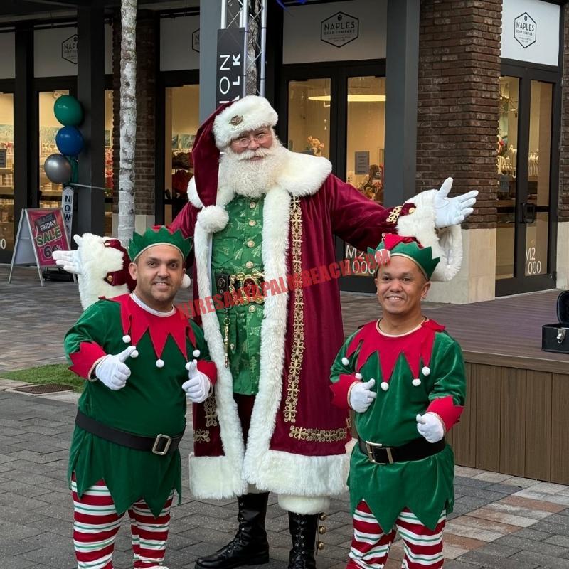 Santa Claus impersonator in a burgundy coat with two elves giving thumbs-up outside a storefront.